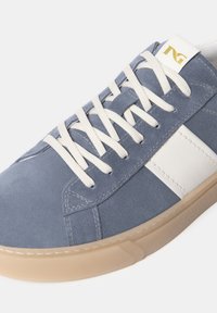 Zapatillas de ante azul con acentos de piel blanca, con cordones crema y suela de goma beige. Logo en dorado cerca de la lengüeta.