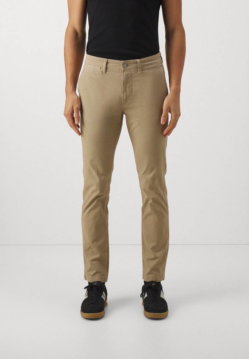 Homme portant un pantalon beige slim, des baskets noires à bandes blanches, et un haut noir sans manches, debout sur un fond blanc uni.