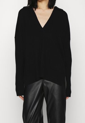 Maglione nero a V, con una vestibilità morbida e maniche lunghe. Presenta dettagli sulle spalle e un orlo posteriore leggermente più lungo. Indossato con pantaloni in pelle nera.