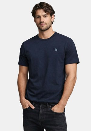 Mann mit kurzem dunklem Haar, der ein marineblaues T-Shirt mit kurzen Ärmeln und schwarze Jeans trägt, steht mit den Händen in den Taschen vor weißem Hintergrund.