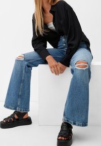 Camicia nera a maniche lunghe sopra una canotta grigia, abbinata a jeans a zampa d'elefante blu chiaro con strappi sulle ginocchia, e sandali neri con piattaforma e fasce.