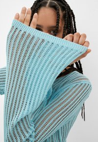 Helles blaues Strickpullover mit losem, offenem Gewebe, verlängerten Ärmeln und einem gerippten Halsausschnitt; der Stoff zeigt ein gestreiftes Muster.