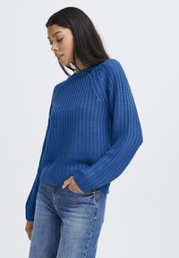 Pull en tricot bleu avec un motif texturé, col rond et manches longues, associé à un jean en denim. Gros plan sur le pull.