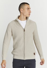 Suéter beige con cremallera, de textura acanalada y cuello alto. Presenta un detalle en piel marrón cerca del dobladillo. Usado sobre una camiseta blanca.