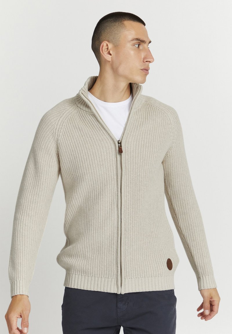 Suéter beige con cremallera, de textura acanalada y cuello alto. Presenta un detalle en piel marrón cerca del dobladillo. Usado sobre una camiseta blanca.