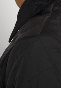 Veste matelassée noire avec un col en velours côtelé, un motif de couture diagonale et une texture lisse et brillante.