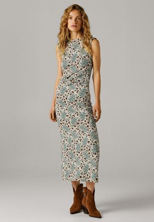 Vrouw met krullend haar, gekleed in een mouwloze midi-jurk met bloemenprint en bruine suède enkellaarsjes, staand tegen een effen achtergrond.