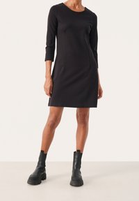 Robe noire ajustée avec manches trois-quarts, col rond et poches latérales. Associée à des bottines noires épaisses avec une semelle en caoutchouc.