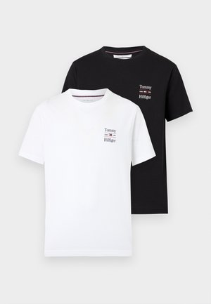 Mężczyzna w beżowej czapce, czarnej koszulce Tommy Hilfiger, niebieskich dżinsach i czarnych butach, stojący z jedną ręką w kieszeni na białym tle.