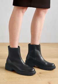 Bottines noires en cuir lisse avec un bout arrondi, semelle en caoutchouc épaisse et des détails de fermeture éclair sur le côté, posées sur un sol en bois.