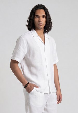 Giovane uomo con capelli castani di media lunghezza che indossa una camicia bianca a maniche corte con bottoni e pantaloni bianchi abbinati, in piedi con una mano in tasca.