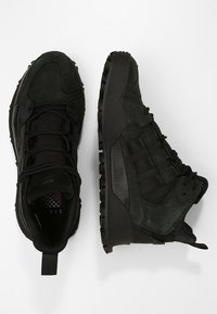 Ett par svarta högskor med texturerade sulor, snörning fram och vadderade kragar, visade mot vit bakgrund.