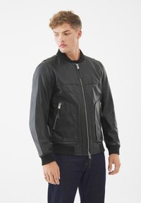 Chaqueta bomber de cuero negro con puños y cuello acanalados, que presenta una cremallera frontal y dos bolsillos laterales con cremallera. Textura suave y diseño ajustado.