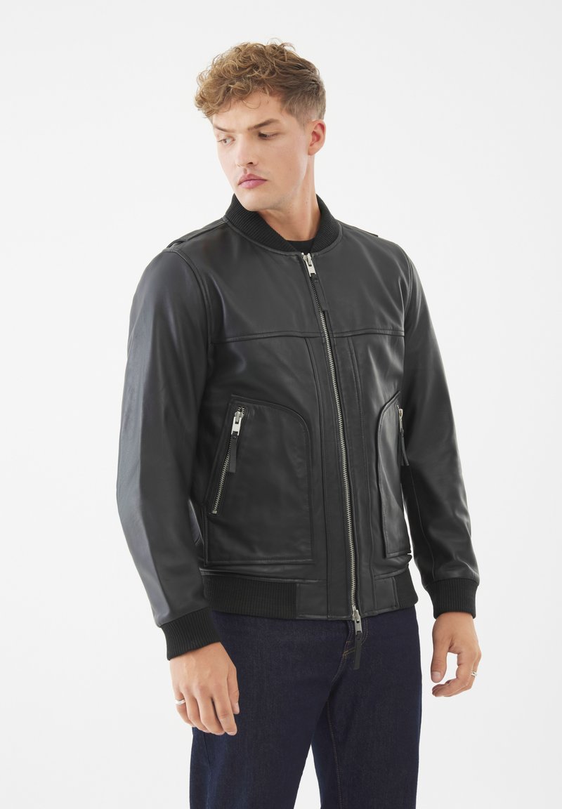 Chaqueta bomber de cuero negro con puños y cuello acanalados, que presenta una cremallera frontal y dos bolsillos laterales con cremallera. Textura suave y diseño ajustado.