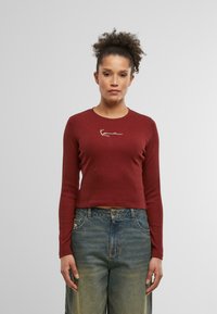 Burgundy long-sleeve top with a cropped design and embroidered white logo at the chest, paired with loose, faded blue denim jeans.
