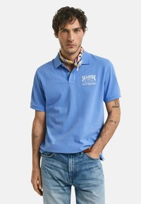 Ljusblå pikétröja med krage och en logotyp med texten "Seaside" på vänstra bröstet, ihop med ljusblå denimjeans och en mönstrad scarf.