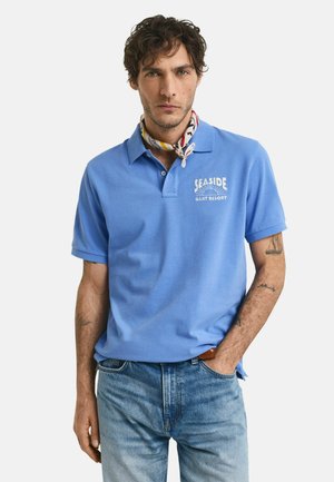 Lyseblå polo-shirt med krave og et logo med teksten "Seaside" på venstre bryst, parret med lyseblå denimjeans og et mønstret tørklæde.