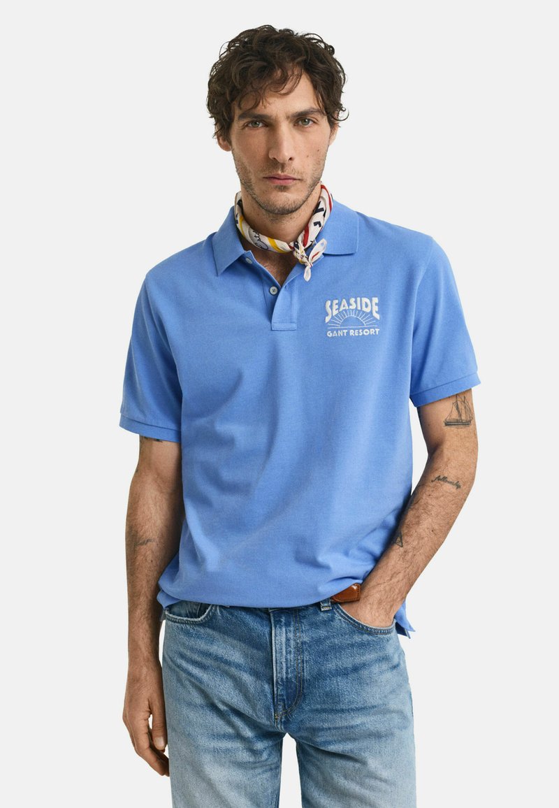 Ljusblå pikétröja med krage och en logotyp med texten "Seaside" på vänstra bröstet, ihop med ljusblå denimjeans och en mönstrad scarf.