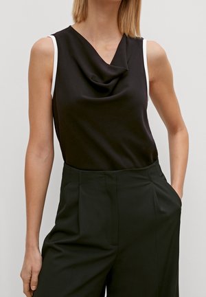 Femme portant un haut noir sans manches à encolure drapée et un pantalon noir taille haute, une main dans la poche, sur un fond uni.