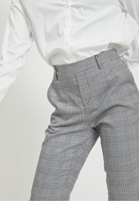 Spodnie w szary i biały wzór w kratkę, o dopasowanym kroju, wysokim stanie i gładkim materiale. Posiadają szlufki na pasek i eleganckie boczne kieszenie.