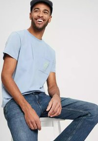 Jasnoniebieska bawełniana koszulka z kieszenią na piersi i zielonym wzorem graficznym. Połączona z niebieskimi dżinsami, siedząca na stołku.