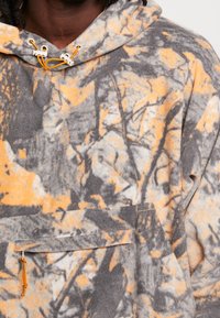 Fleece hoodie i grå med et orange og sort træmønster, der har en kængurulomme og justerbare snore i hætten.