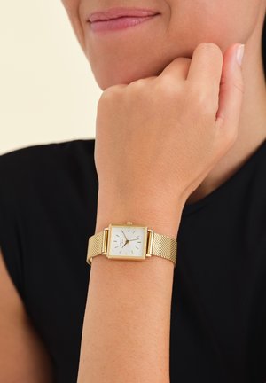 Montre avec bracelet en maille dorée, dotée d'un cadran carré blanc, de marqueurs d'heure dorés et d'un boîtier rectangulaire fin. Portée au poignet contre une chemise noire.