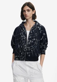 Sudadera con cremallera color navy con patr�ón gráfico abstracto en plata, que cuenta con una capucha con cordón, puños acanalados y bolsillos frontales, combinada con pantalones cargo blancos.