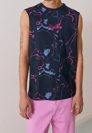 Osoba ubrana w granatową bluzkę bez rękawów z różowo-niebieskimi abstrakcyjnymi wzorami oraz różowe spodnie, stojąca na tle jednolitego tła.