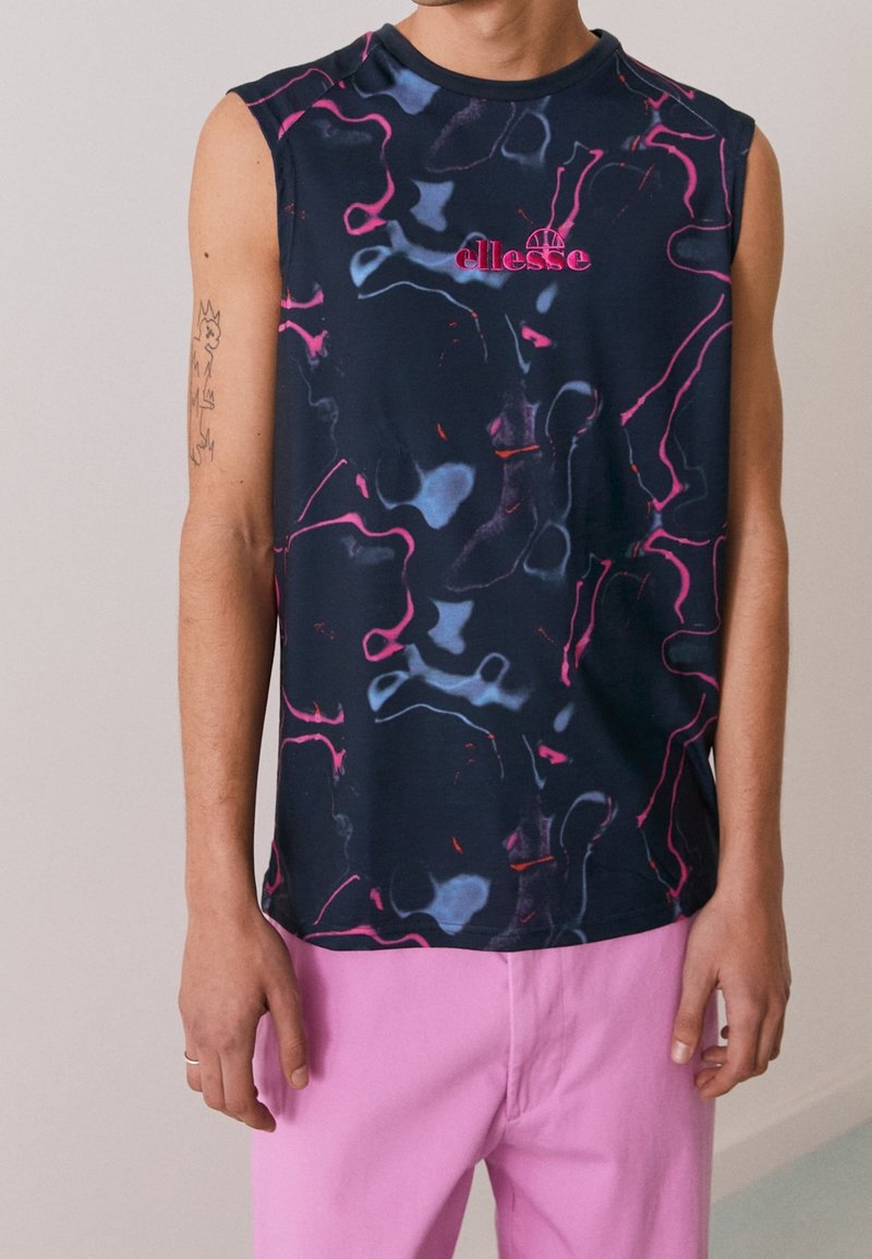 Personne portant un haut sans manches bleu marine avec des motifs abstraits roses et bleus et un pantalon rose, debout devant un fond uni.