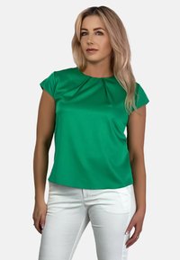 Zielona bluzka z krótkim rękawem, okrągłym dekoltem i plisowanym detalem z przodu, zestawiona z białymi spodniami. Gładki, lekki materiał.