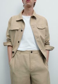 Giacca in lino beige con maniche arrotolate, due tasche sul petto e bottoni neri, abbinata a una maglietta bianca e pantaloni beige.