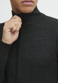 Svart texturerad zip-up tröja med hög ribbad krage, med ett subtilt honungskaksmönster, halvdraget, med en närbild av halsringningen.