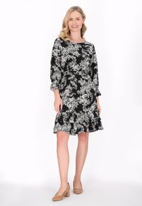 Schwarzes Blumenkleid mit lässigem Schnitt, Dreiviertelärmeln, einem fransigen Saum und weißen Blumendrucken. Kombiniert mit beigen Blockabsätzen.