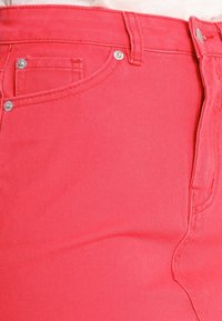 Jean en denim rose corail avec une texture lisse, un design à cinq poches et des accents en métal argenté. Détails cousus le long des coutures.