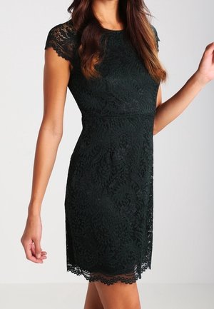 Frau trägt ein schwarzes, figurbetontes Kleid mit kurzen Ärmeln und floralen Spitzenmustern, steht vor einem schlichten hellen Hintergrund.