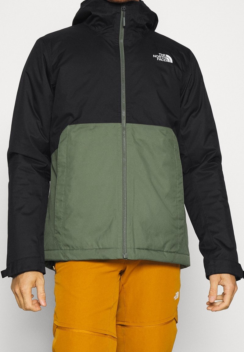 Homme portant une veste à capuche noire et verte zippée et un pantalon jaune moutarde, debout les bras détendus sur les côtés.
