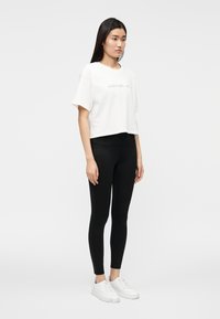 T-shirt branca cropped com texto da marca em relevo, combinada com leggings pretas justas e ténis brancos. Tecido suave e design minimalista.