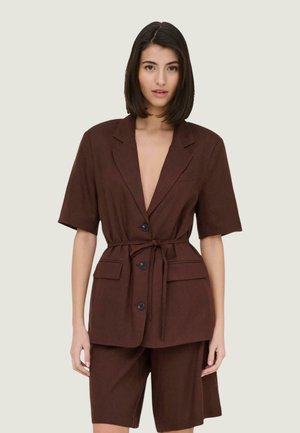 Vrouw met kort donker haar draagt een bruine blazer met ceintuur en knopen en bijpassende knielange shorts tegen een effen achtergrond.