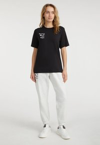 Sort bomulds T-shirt med korte ærmer og hvid tekst, parret med grå sweatpants med sort tekst på venstre ben og hvide sneakers.