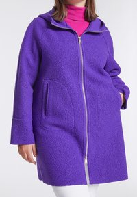 Manteau polaire violet avec capuche, surface texturée, fermeture éclair complète, deux poches avant et manches longues, porté par-dessus un col roulé rose.