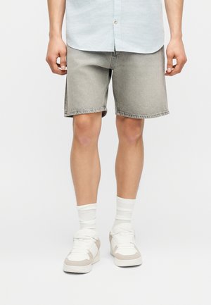 Homme portant un short en denim gris clair, des chaussettes blanches montantes et des baskets blanches, les mains détendues le long du corps sur un fond uni.