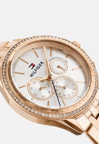 Reloj de oro rosado con esfera blanca, acentos de cristal, tres subesferas y marcadores de horas en tono dorado. Presenta un diseño de pulsera de eslabones.