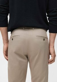 Beige Stoffhosen mit glatter Textur, ausgestattet mit zwei Gesäßtaschen mit Knopfverschlüssen, getragen mit einem schwarzen Pullover.