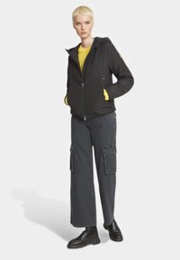 Giacca nera con zip e cappuccio, abbinata a un maglione giallo e pantaloni cargo grigi. Presenta tasche e dettagli in tessuto strutturato.