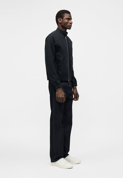 Άνδρας που στέκεται πλάγια φορώντας μαύρο μπουφάν με φερμουάρ, μαύρο παντελόνι και λευκά αθλητικά παπούτσια σε απλό λευκό φόντο.