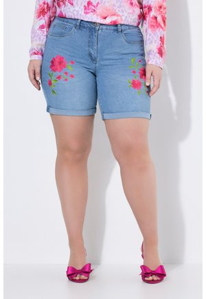 Person iført blå denimshorts med pink blomstret broderi og pink peep-toe hæle med store sløjfer, stående mod en hvid baggrund.