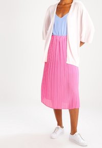 Růžová skládací sukně střední délky, doplněná světle modrým topem a bílým svetrem. Celkový outfit doplňují bílé tenisky.