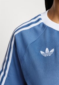 Haut de sport bleu en tissu texturé. Présente des accents à trois bandes blanches et un petit logo blanc brodé sur la poitrine.