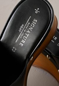 Sandali slide in pelle nera con base in legno marrone, dotati di dettagli in metallo argentato, marchio stampato sulla soletta e una texture liscia.
